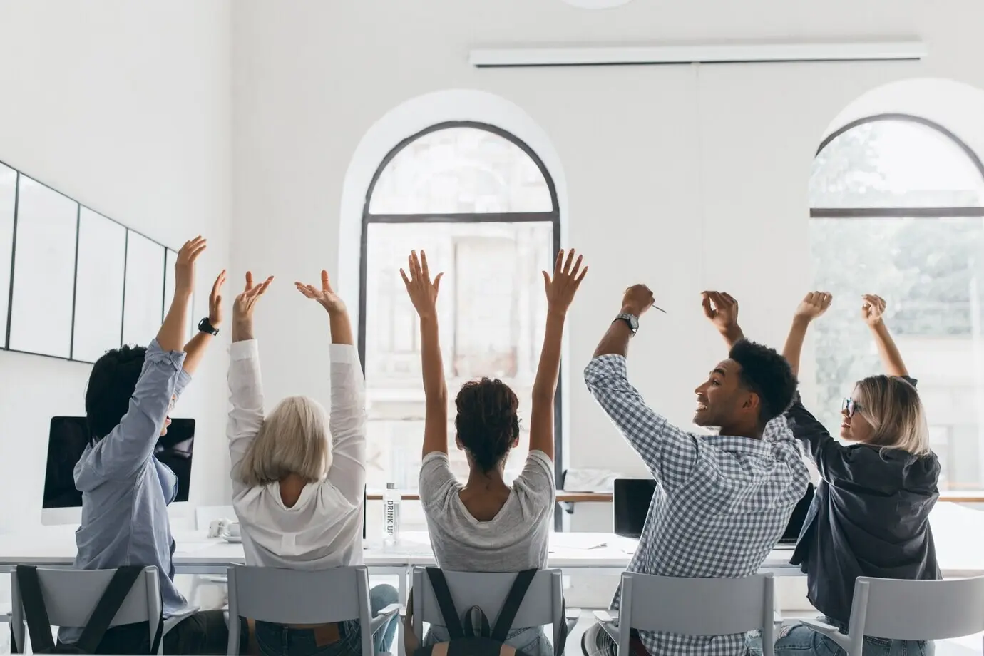 Frau mit blondem Haar im formellen Hemd, die mit den Händen winkt, sitzt zwischen Kollegen in einem großen, hellen Konferenzsaal. Foto von hinten: müde Manager strecken sich während einer Besprechung im Büro.
